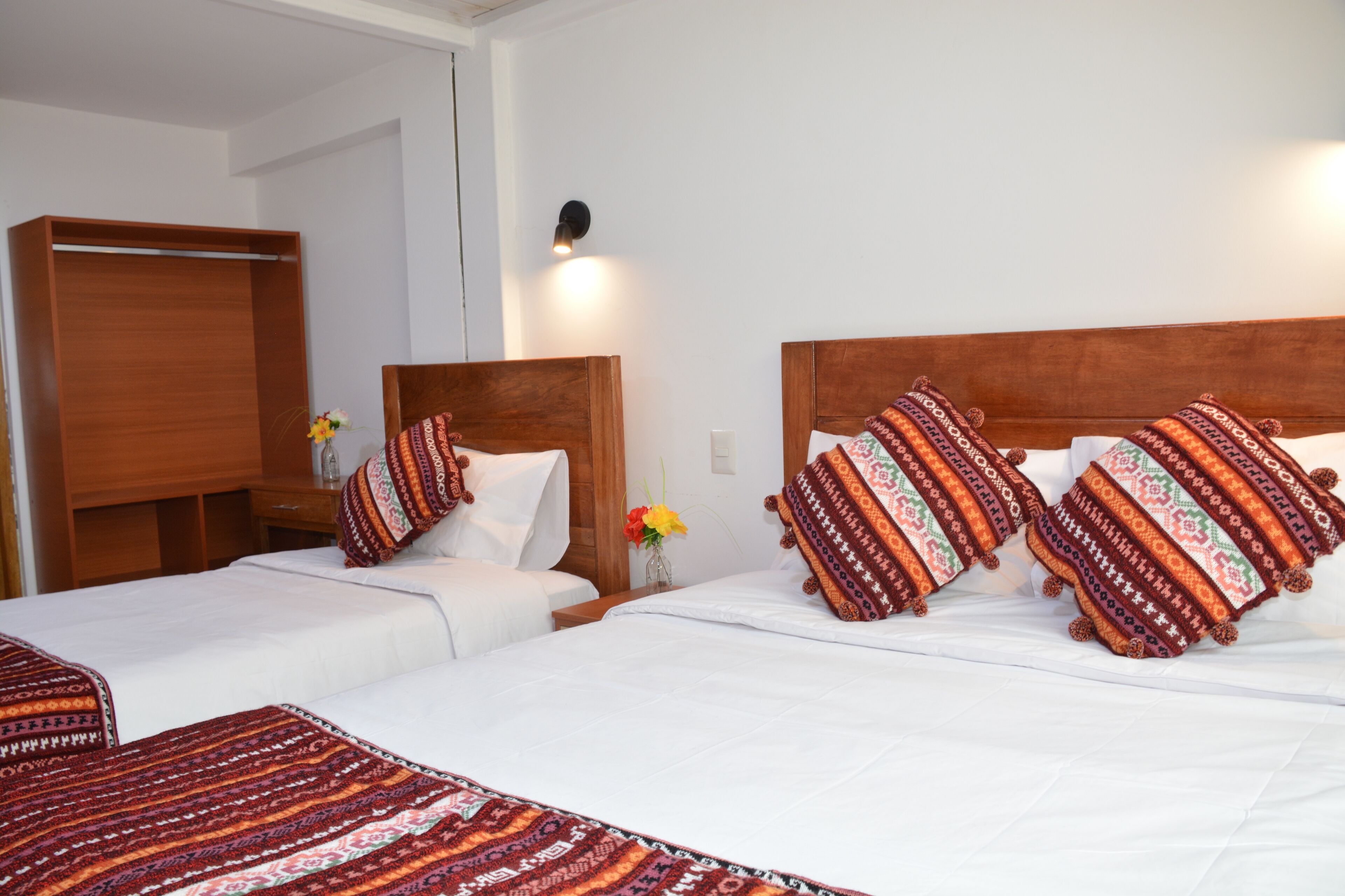 Habitación de hotel con dos camas, cada una con almohadones estampados y cabeceros de madera. Hay una mesita de noche con un jarrón de flores y una lámpara fijada a la pared.