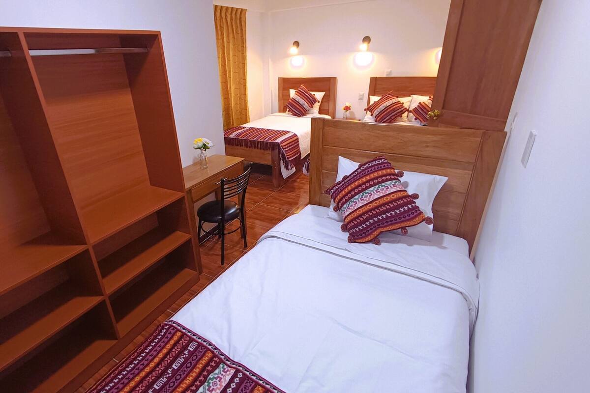 A hotel room with a large wooden wardrobe, a desk with a chair, two beds with patterned pillows, and a mirror above the bed.