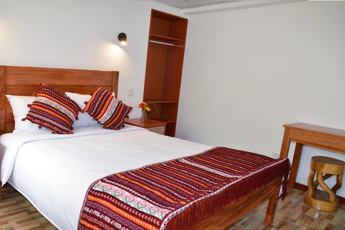 A neatly made bed with patterned pillows, a wooden headboard, a nightstand with a vase of flowers, and a wooden desk with a chair.