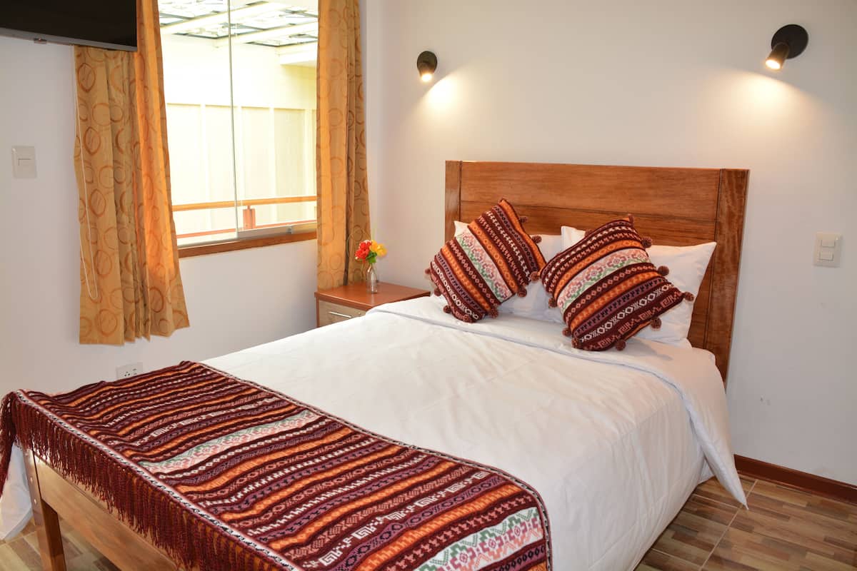A neatly made bed with patterned pillows, a bedside table with a vase of flowers, and a window with curtains.