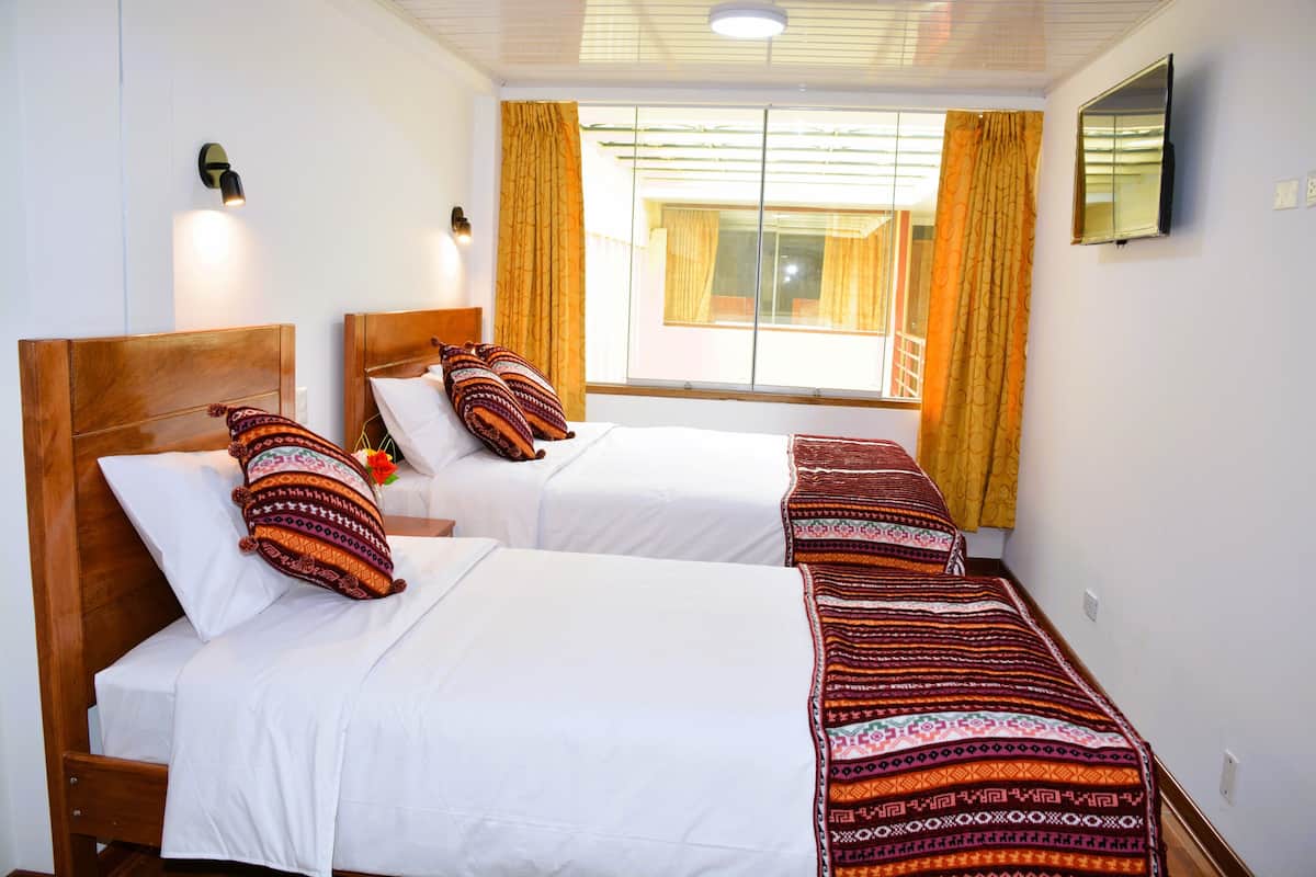 A hotel room with two single beds, a large window with curtains, a flat-screen TV mounted on the wall, and a ceiling light fixture.