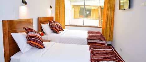 A hotel room with two single beds, a large window with curtains, a flat-screen TV mounted on the wall, and a ceiling light fixture.
