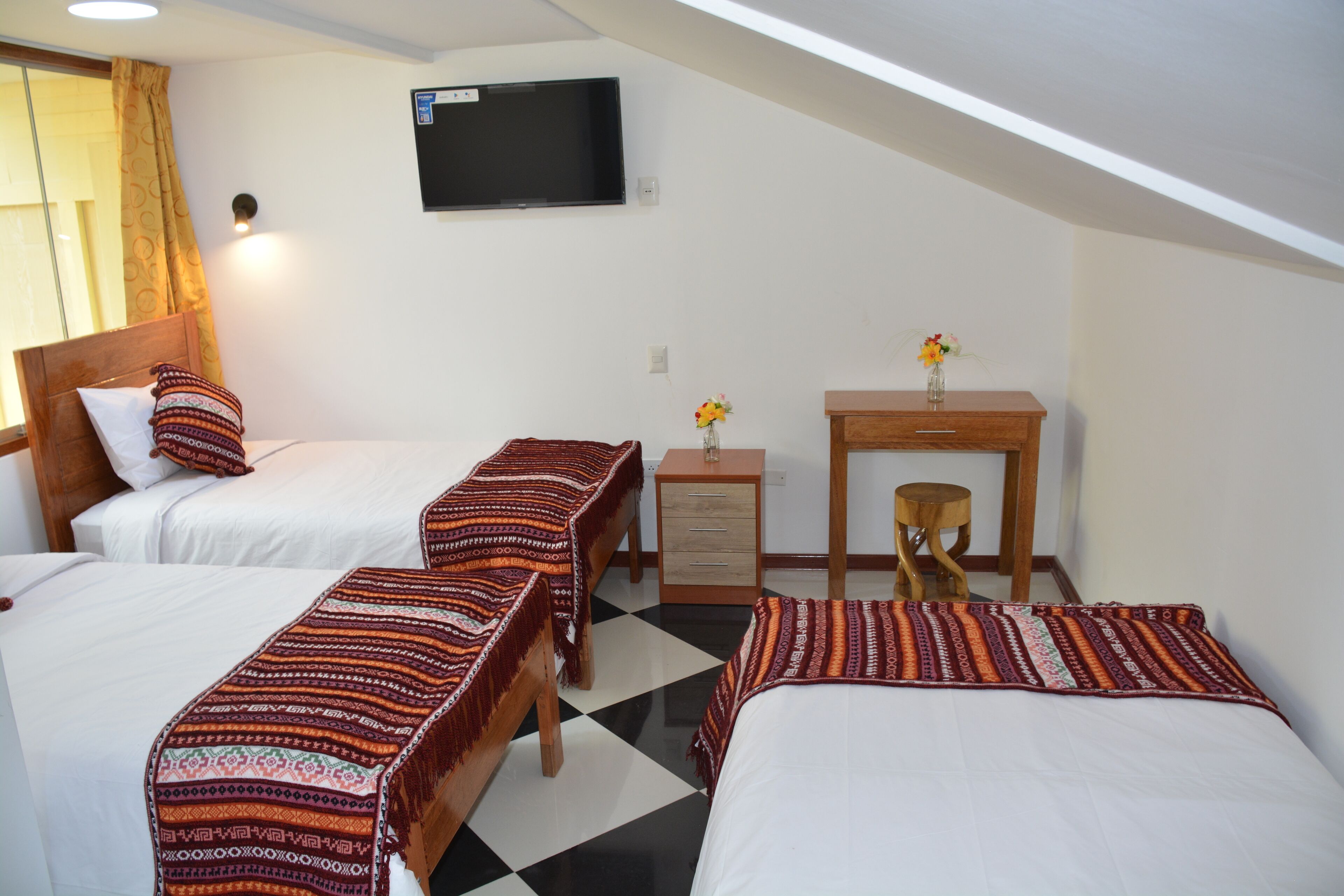 A hotel room with two beds, a desk, a chair, a TV, and a small table with flowers.