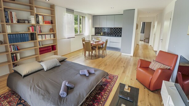 Een moderne hotelkamer met een groot bed, een zithoek met een rode fauteuil, een boekenkast, en een keukenruimte zichtbaar door een open deur.