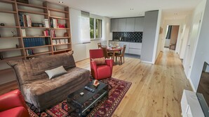Een moderne woonkamer met een bank, een rode fauteuil en een salontafel. Er is een boekenplank vol met boeken en een keuken in de achtergrond.