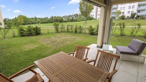 Een overdekt terras met een houten tafel en stoelen, een comfortabele lounge en uitzicht op een gazon met bomen en gebouwen op de achtergrond.