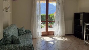 Quarto com sofá, uma estante preta e porta que dá para uma varanda com vista para vegetação e montanhas.