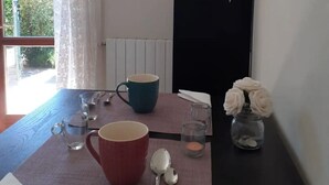 Uma mesa de jantar com um jogo americano, uma caneca vermelha, uma caneca azul, uma garrafa de vinho, um vaso com flores brancas e um conjunto pequeno de colheres de metal.