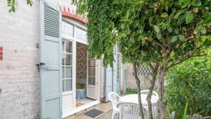 Une petite terrasse extérieure avec une table et des chaises blanches, un pot de fleurs bleu et une porte donnant sur une pièce dont les murs sont recouverts d’un papier peint à motifs.
