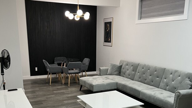 A modern living room with a sectional sofa, a white coffee table, and a dining area with a glass table and chairs. - Modern Private Suite in a serene location 15 minutes away from the Airport. (Airdrie)