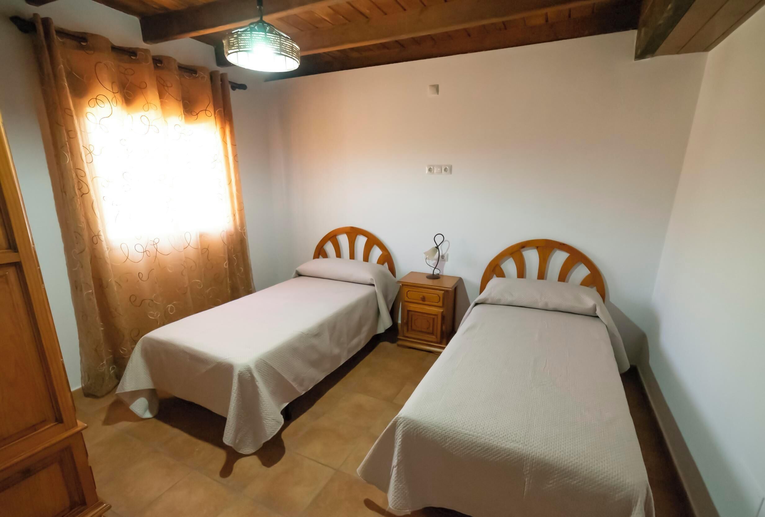 A room with two single beds, wooden headboards, a nightstand with a lamp, and a window with curtains.