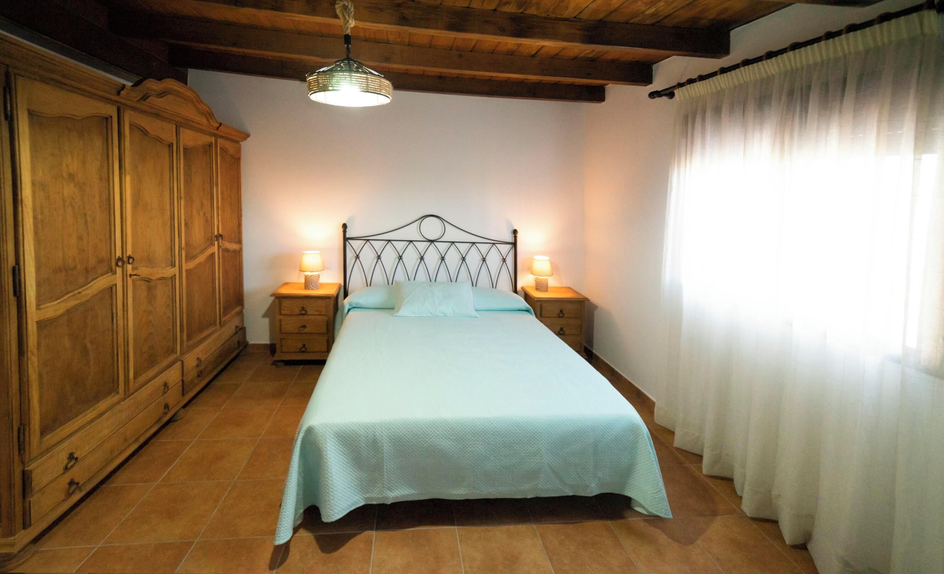 Une chambre à coucher comprenant un lit, une armoire en bois, deux tables de chevet avec des lampes, un luminaire suspendu et une fenêtre avec des rideaux.