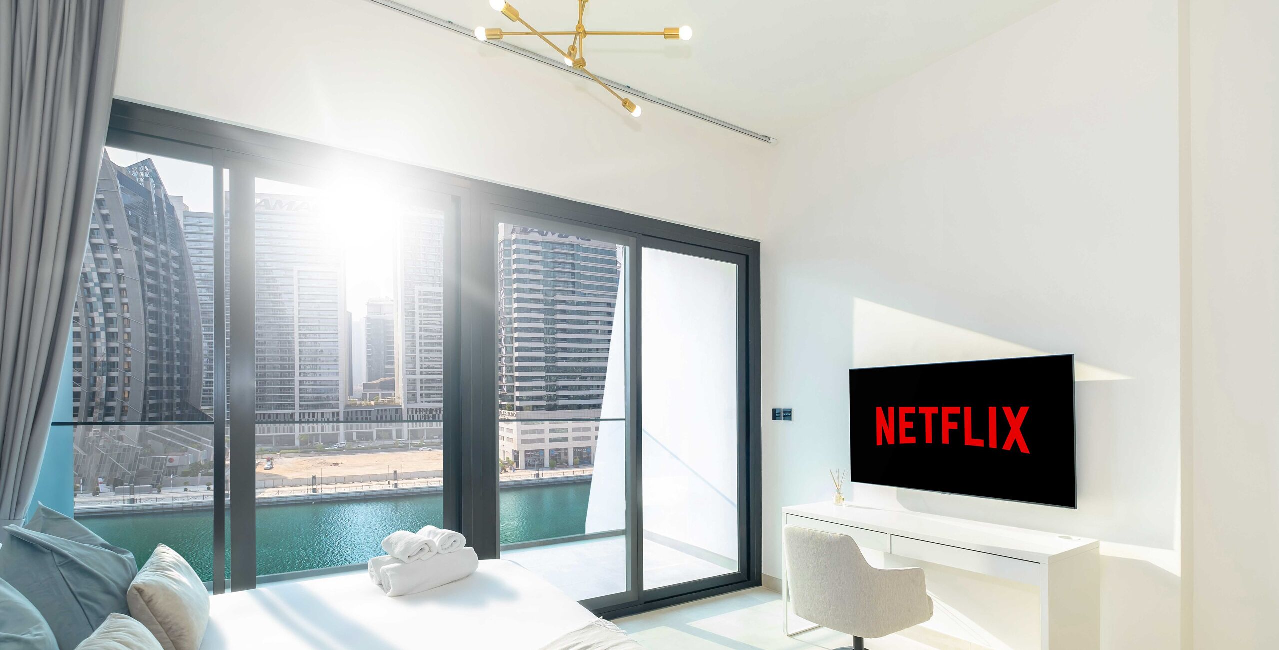 A modern hotel room with a large window offering a view of a cityscape and water. A flat-screen TV mounted on the wall displays the Netflix logo.