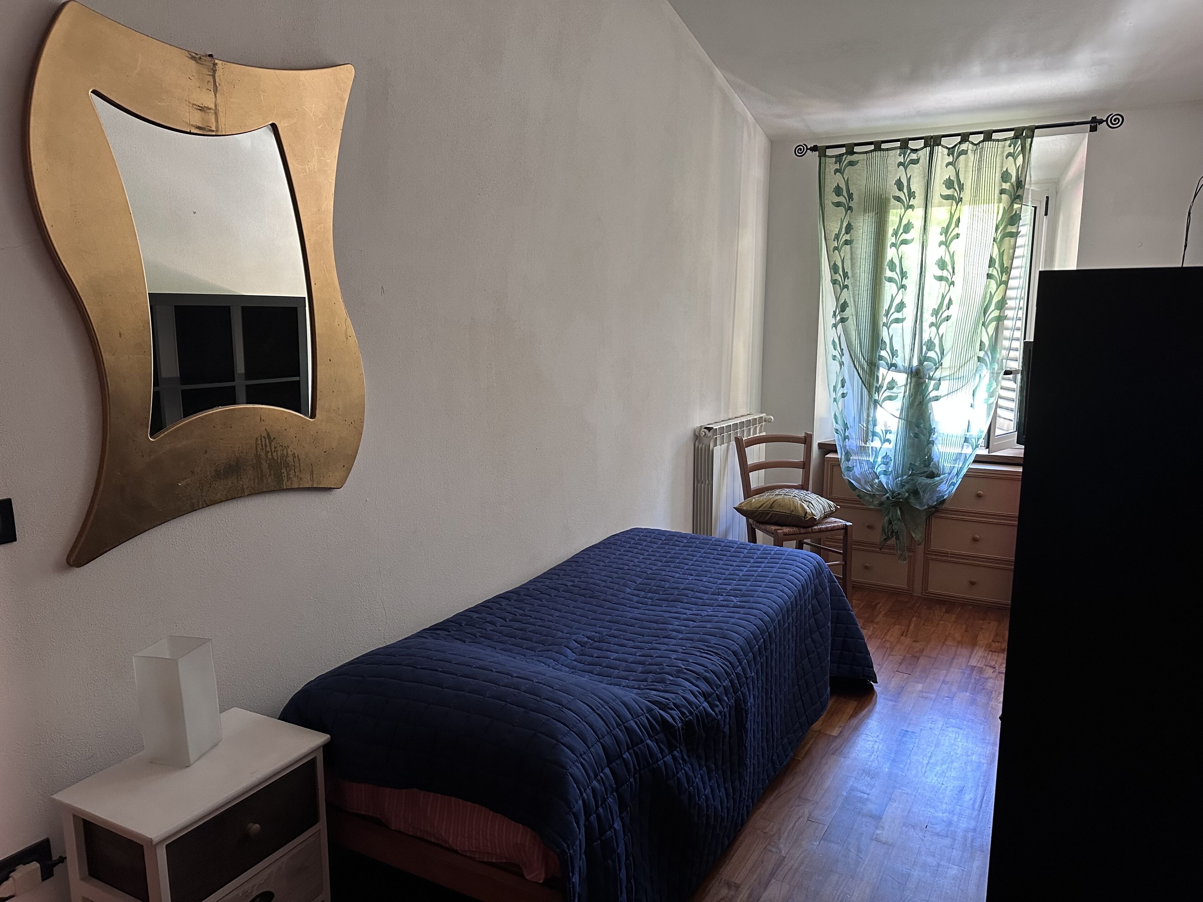 Ein Schlafzimmer mit einem Bett, einem Schminktisch, einem Sessel, einem Heizkörper und einem Fenster mit Vorhängen.