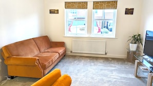 Una sala de estar con un sofá marrón, un sillón amarillo, una mesa de centro de vidrio, un televisor y una ventana que ofrece vistas de un edificio y coches estacionados.