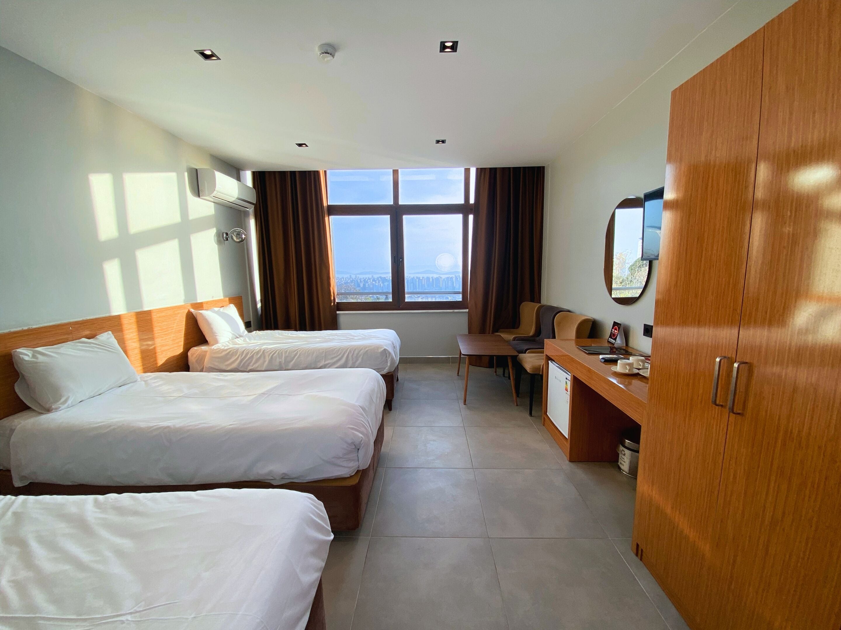 A hotel room with two beds, a desk, a chair, a wardrobe, and a window with a city view.