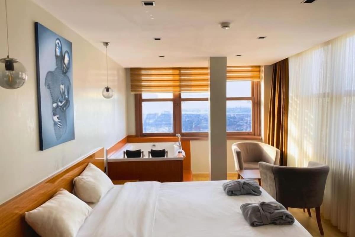 Habitación de hotel con cama, escritorio, silla, ventana con vista a la ciudad y un cuadro en la pared.