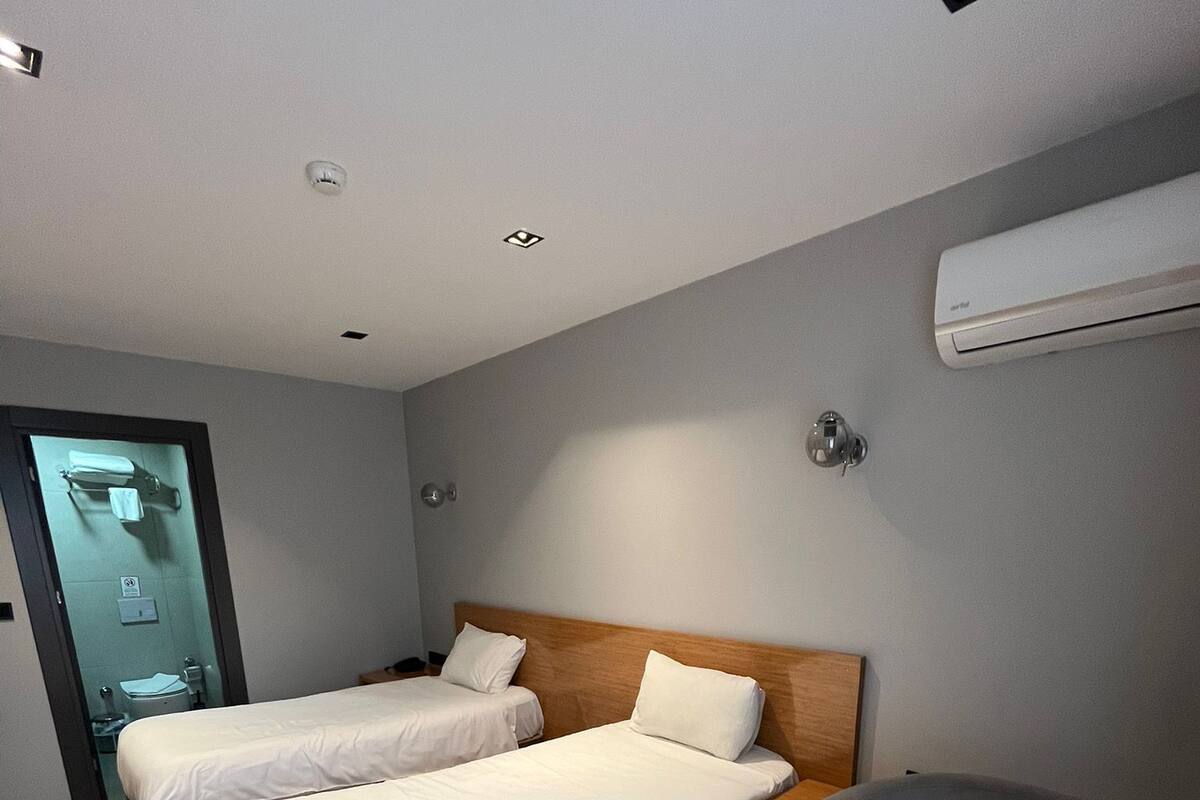 A hotel room with two single beds, a wooden desk, a grey armchair, and an air conditioning unit on the wall.