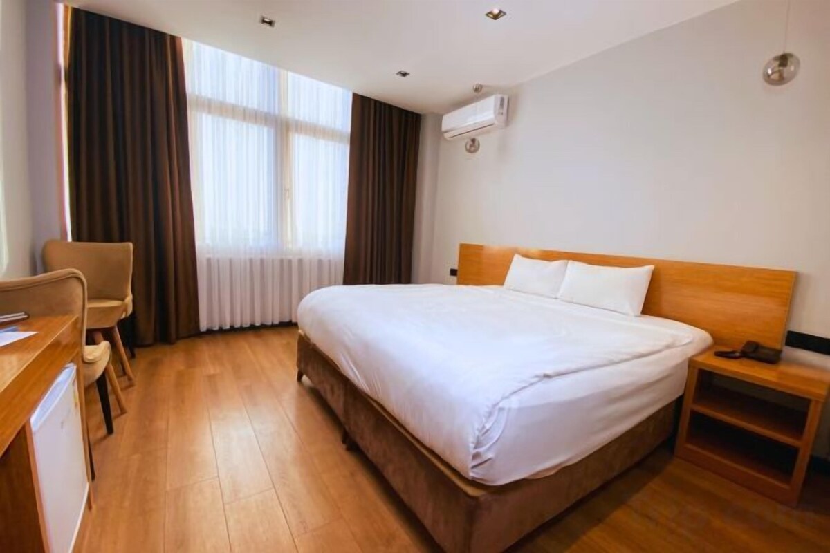 A hotel room with a bed, a desk, a chair, a nightstand, and a window with curtains.