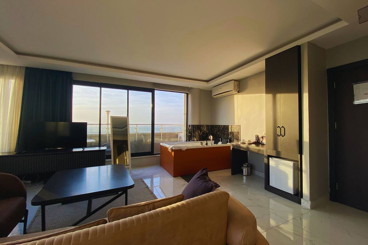 A modern hotel room with a large window, a flat-screen TV, a sofa, a dining table, and a kitchen area.