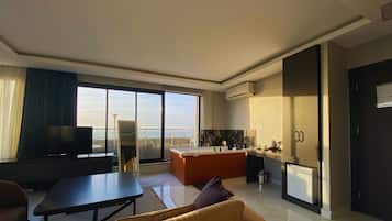 A modern hotel room with a large window, a flat-screen TV, a sofa, a dining table, and a kitchen area.