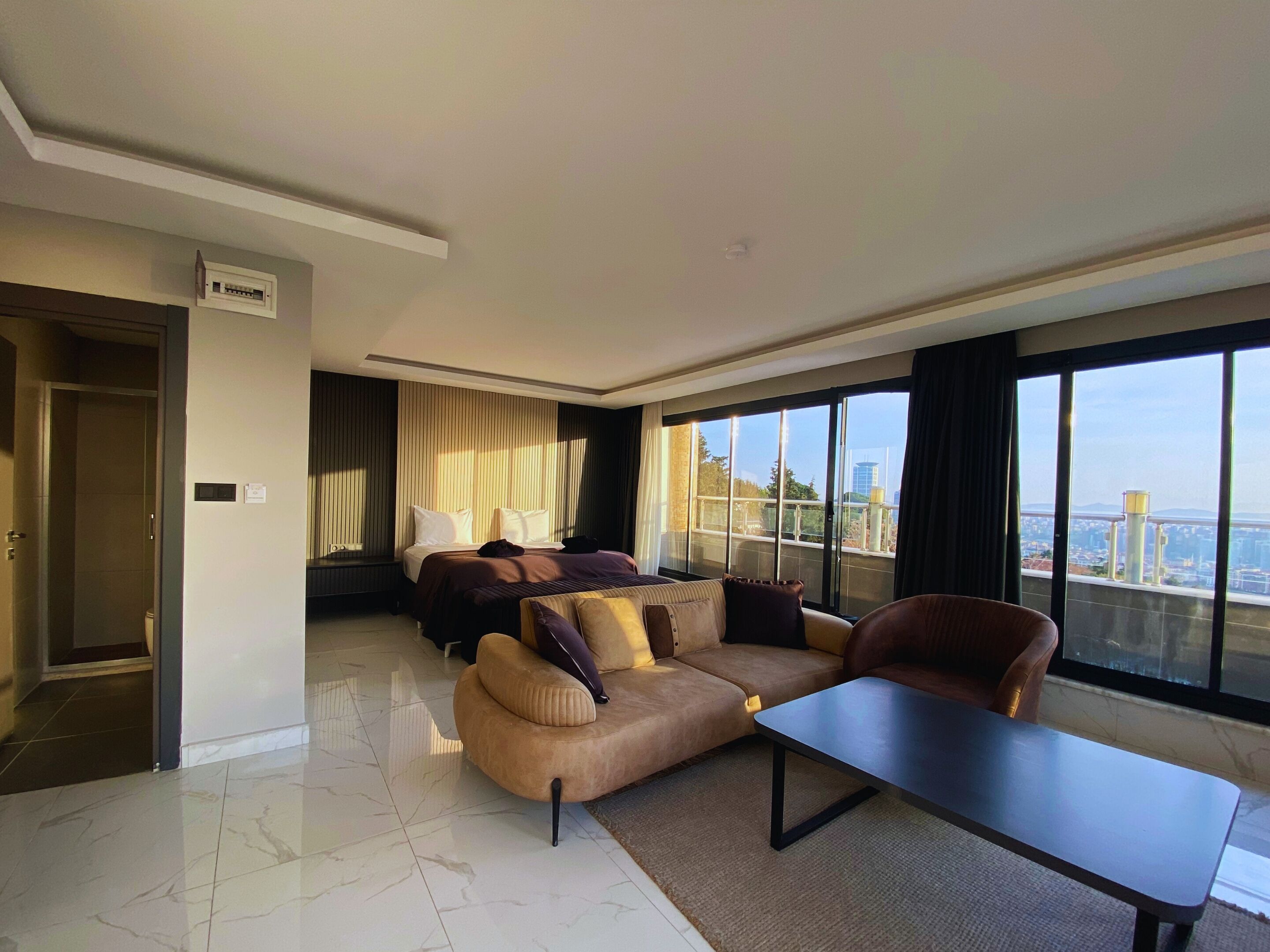 A modern hotel room with a large bed, a sofa, a coffee table, and a view of the city through large windows.