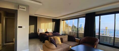 A modern hotel room with a large bed, a sofa, a coffee table, and a view of the city through large windows.