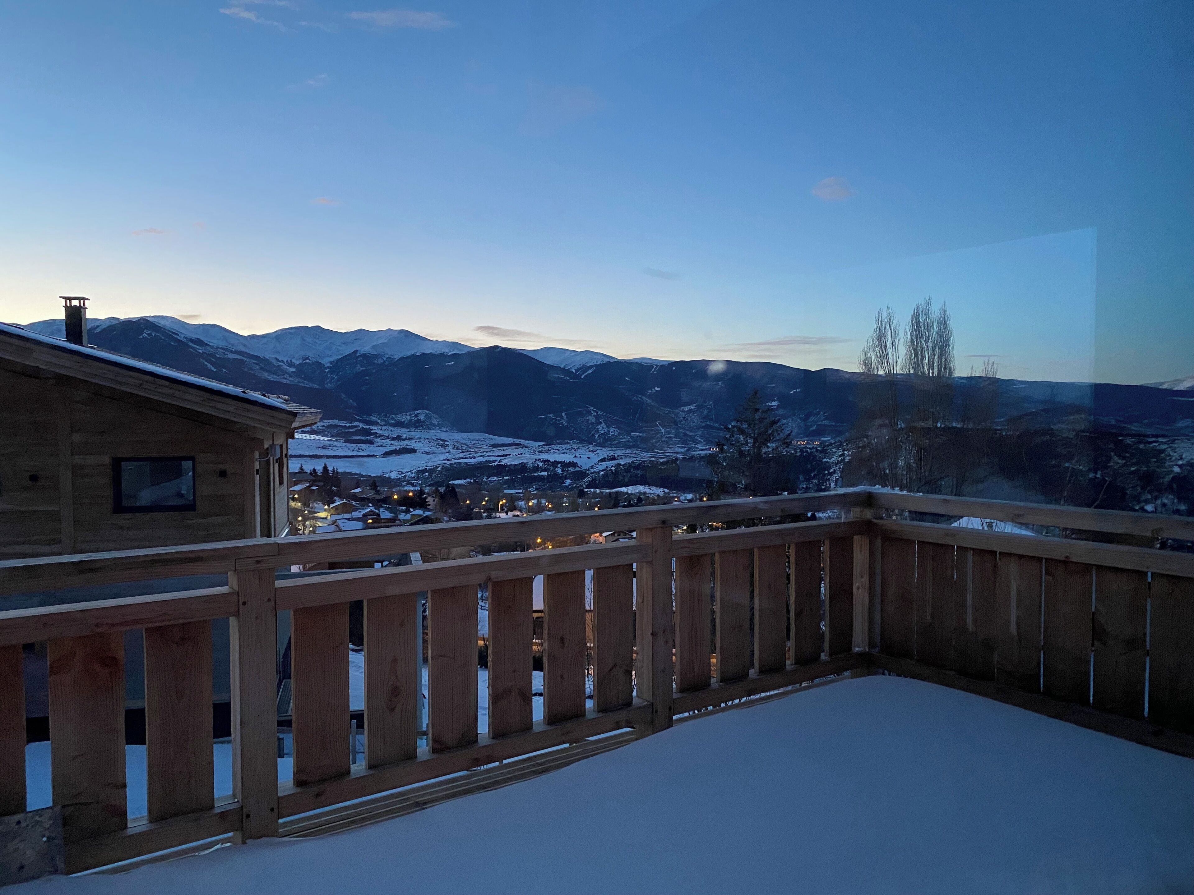 A wooden balcony with a snow-covered ground, overlooking a mountainous landscape with snow-covered houses and a clear sky.