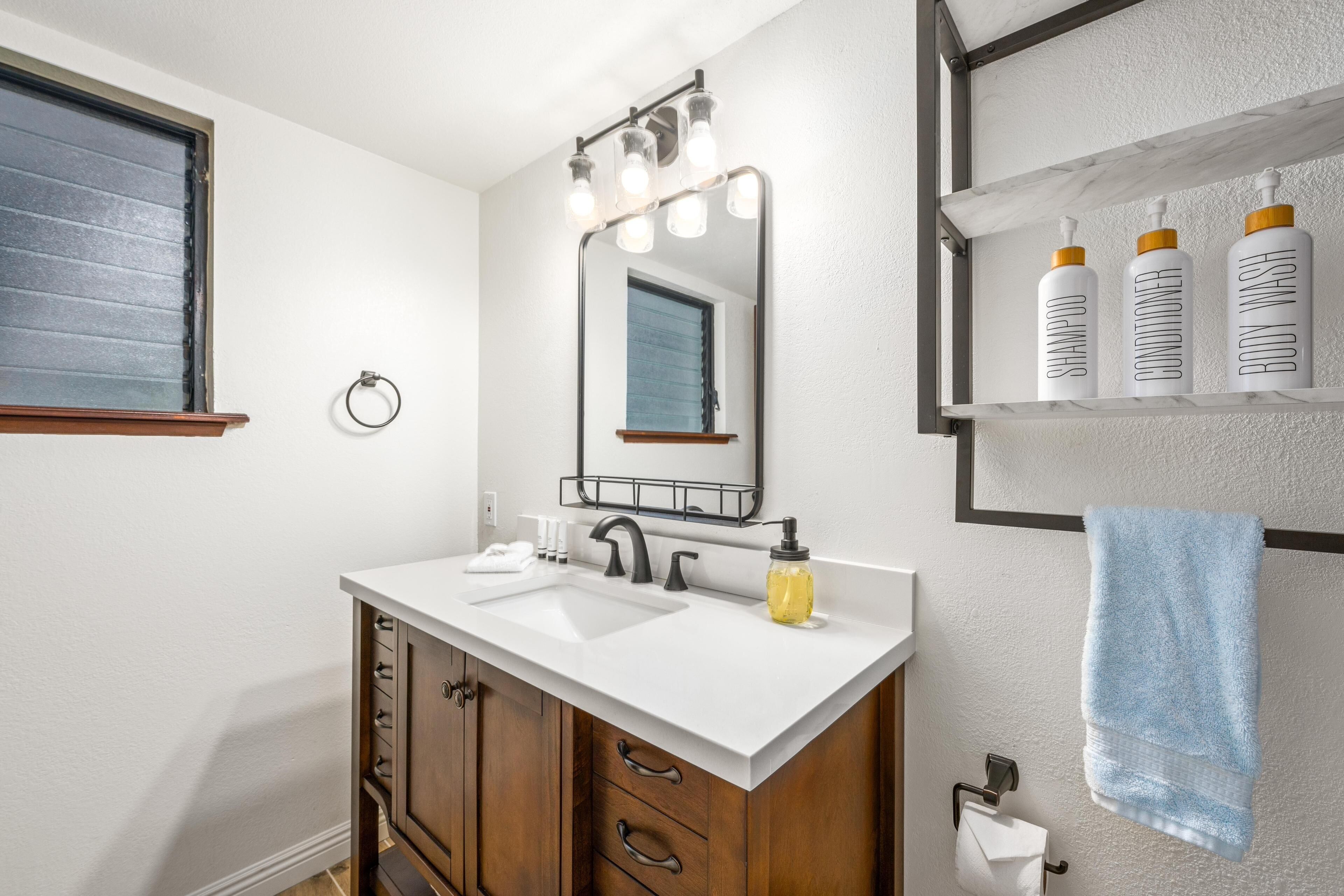 A bathroom with a white vanity, a mirror with lights, a towel rack, and a shelf with toiletries.