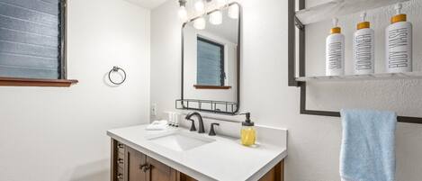 A bathroom with a white vanity, a mirror with lights, a towel rack, and a shelf with toiletries.