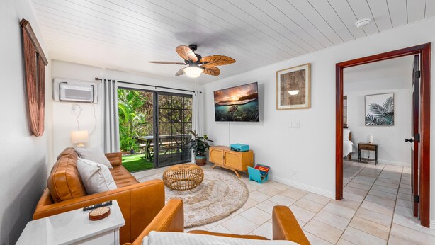A modern living room with a brown leather sofa, a white coffee table, and a ceiling fan. There is a large window with a view of greenery outside.