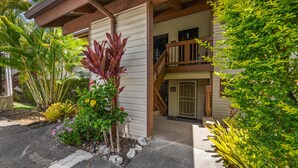 A two-story building with a wooden balcony, a covered entrance, and tropical plants.