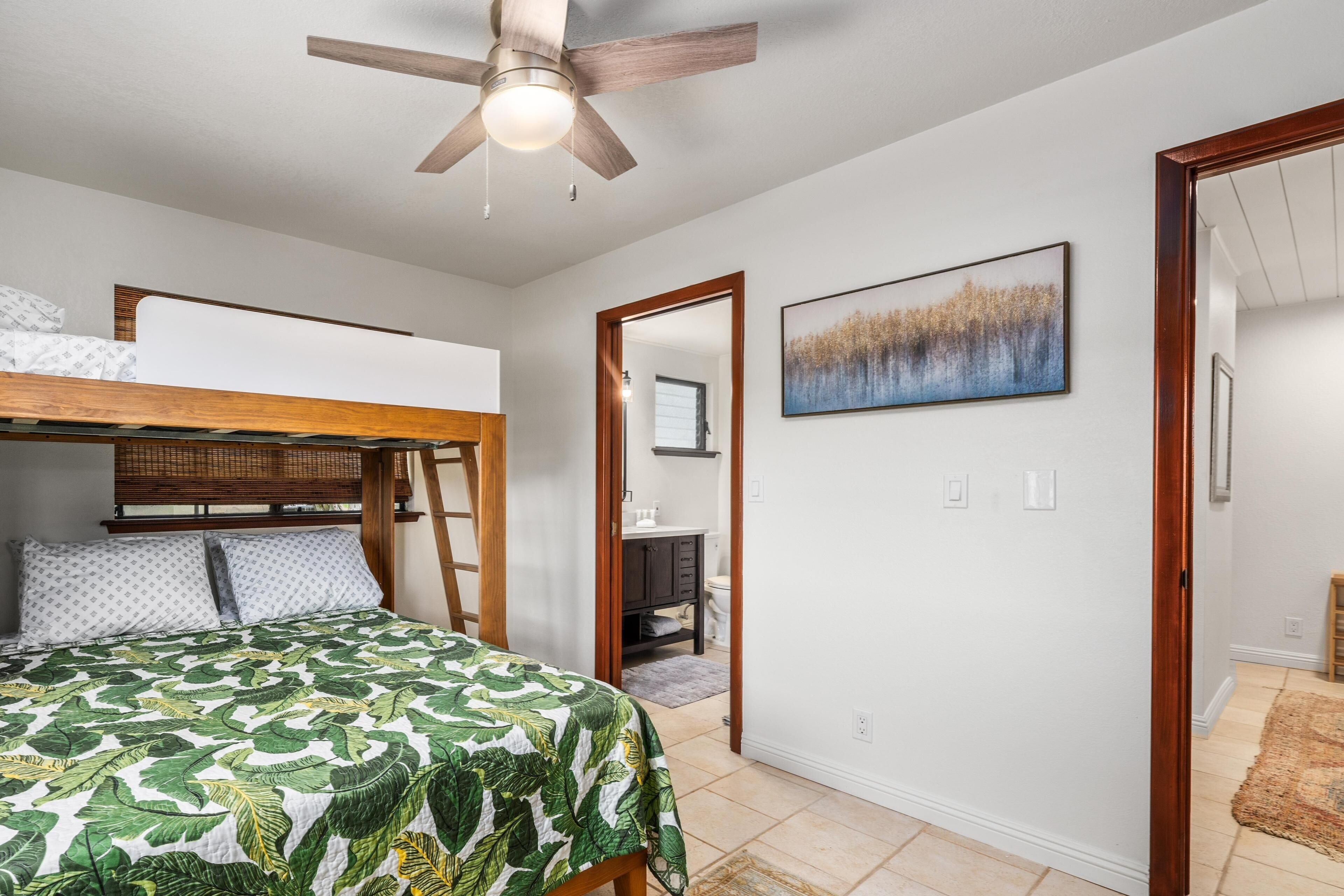 A bedroom with a bunk bed, a ceiling fan, a painting, and a bathroom visible through an open door.