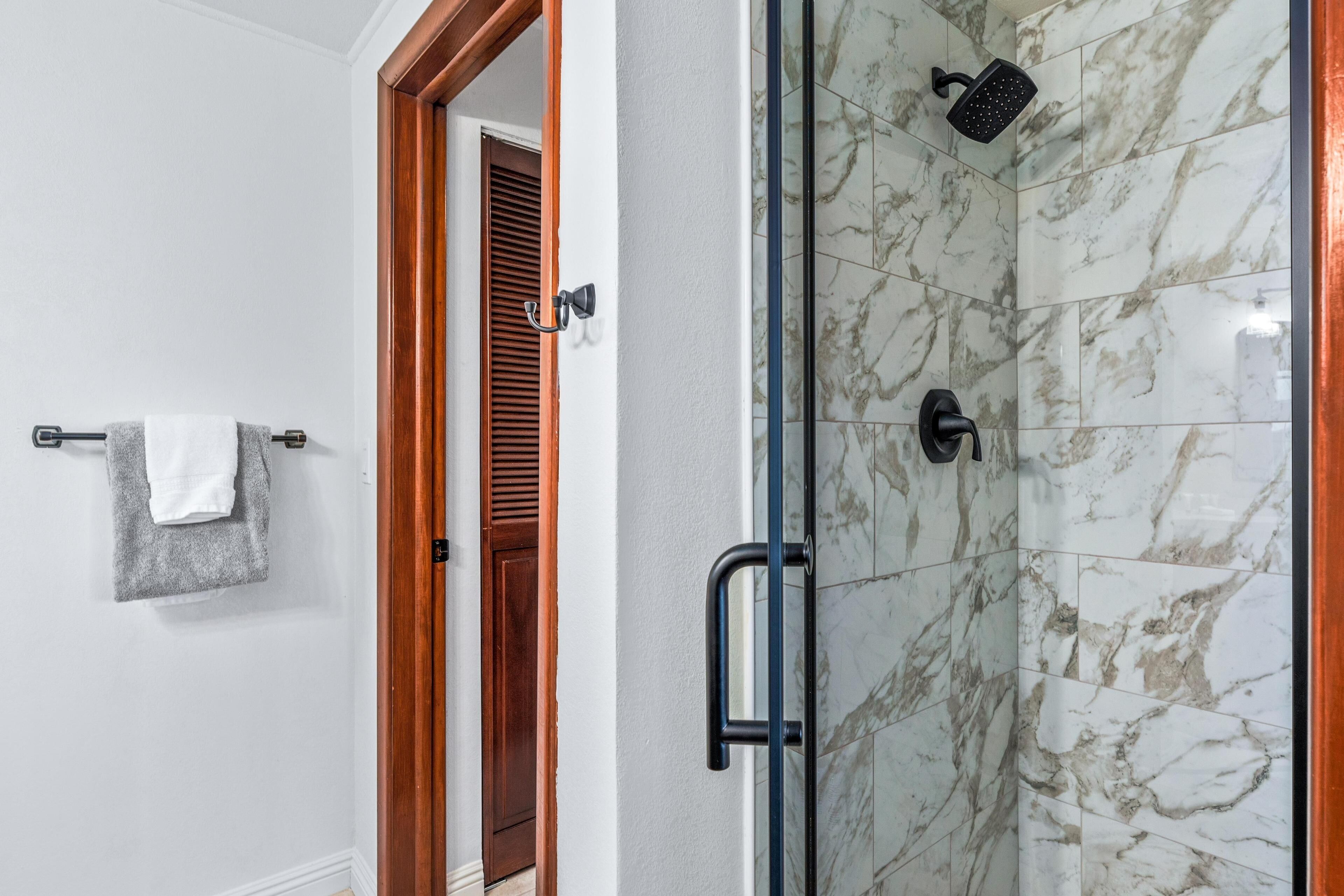 A bathroom with a glass shower enclosure, a wooden door, and a towel rack with two towels.