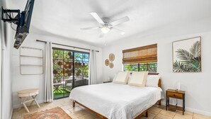 A bedroom with a bed, a ceiling fan, a small table, a nightstand, a rug, and a window with a view of greenery.