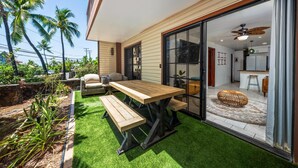 A patio with a wooden table and benches, surrounded by artificial grass and palm trees.