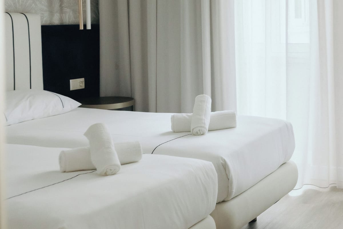 Een moderne hotelkamer met twee bedden, witte lakens, een donker hoofdeinde en een groot raam met dunne gordijnen.
