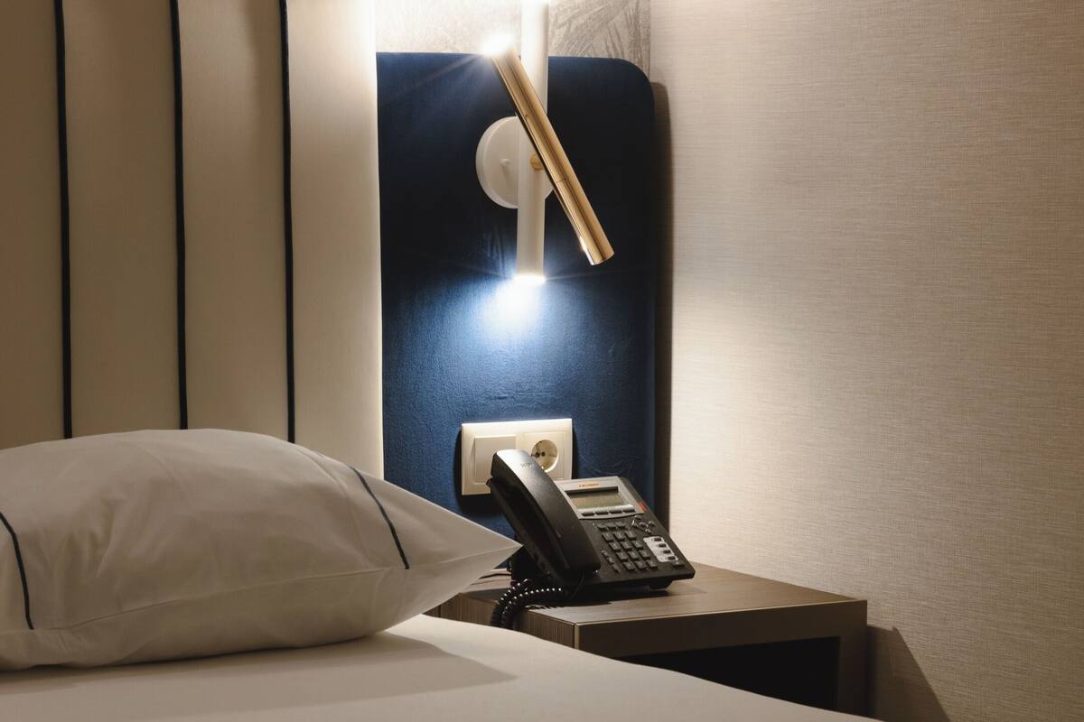 Een keurig opgemaakt bed met een kussen en opgerolde handdoeken, een telefoon op het nachtkastje en een wandlamp.