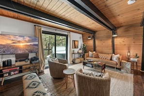 A cozy living room with a sofa, armchairs, a coffee table, and a TV. There is a large window with curtains, a fireplace, and a wooden ceiling.