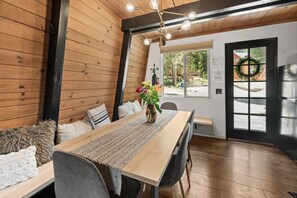 Un interior de madera con una mesa de comedor, sillas, un camino de mesa tejido y un jarrón con flores.