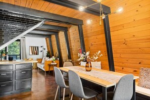 A modern loft-style room with exposed wooden beams, a dining area with a wooden table and chairs, a kitchen with dark cabinetry, and a staircase leading to an upper level. - 2BR Mid Century Retreat • EV, Hot Tub (Big Bear Lake)