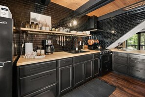 Une cuisine avec des meubles de couleur noire, un plan de travail en bois, une machine à café et des ustensiles suspendus.