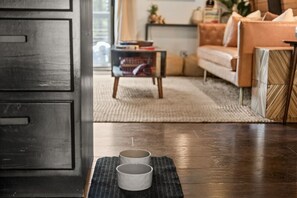 A room with a dark wooden floor, a small table with two white cups, a sofa, and a bookshelf in the background.