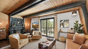 A cozy living room with a stone fireplace, wooden beams, a large window, and a view of the outdoors. - 2BR Retreat: EV, Hot Tub, BBQ & Dog-Friendly (Big Bear Lake)