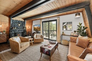 A cozy living room with a stone fireplace, wooden beams, a large window, and a view of the outdoors. - 2BR Retreat: EV, Hot Tub, BBQ & Dog-Friendly (Big Bear Lake)