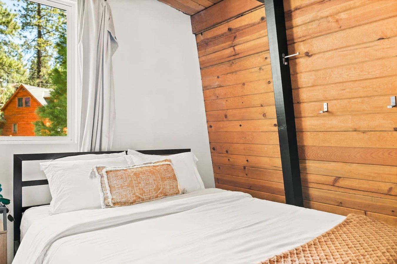 Un dormitorio con una cama, ropa de cama blanca, una pared de madera y una ventana con vista a una casa.