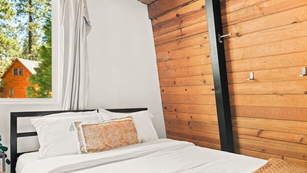 A bedroom with a bed, white bedding, a wooden wall, and a window with a view of a house. - 2BR Retreat: EV, Hot Tub, BBQ & Dog-Friendly (Big Bear Lake)