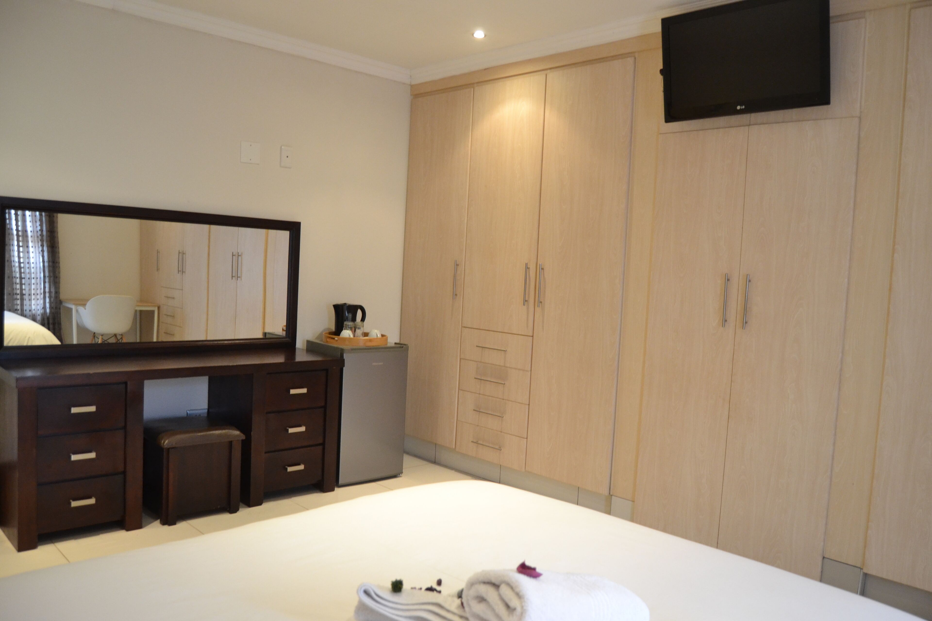 Un dormitorio con una cama grande, un aparador con cajones, un taburete pequeño, un escritorio con una silla, un televisor de pantalla plana montado sobre un armario, y una cafetera sobre el aparador.