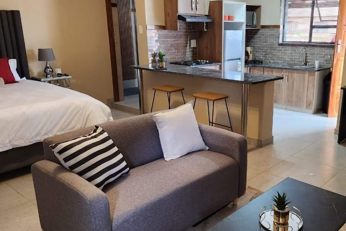 A modern hotel room with a sofa, a small table, a bed, a kitchenette, and a bathroom visible through a door.