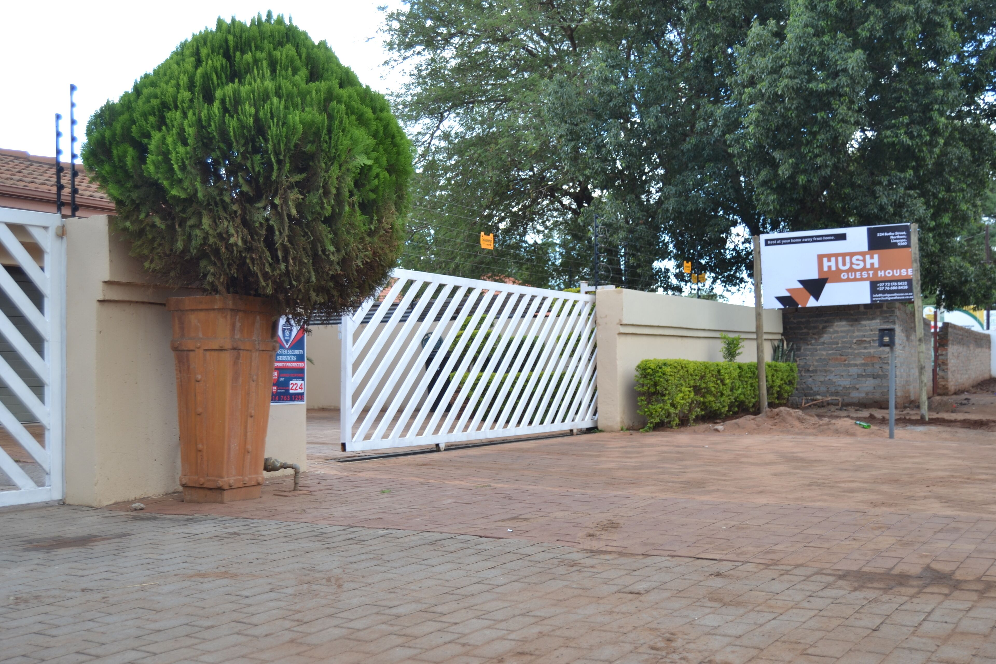A gated entrance with a sign for HUSH GUEST HOUSE, a wooden barrel, and a trimmed bush.